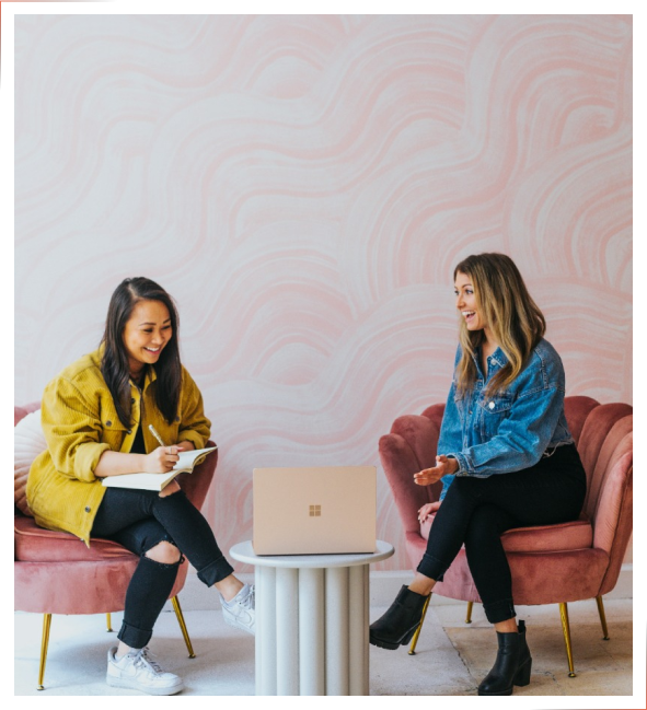 Two women sitting in pink chairs, engrossed in their laptop.